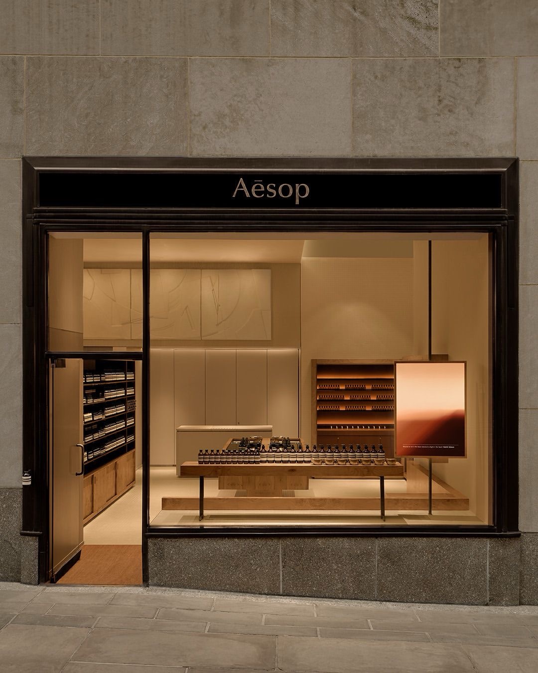 Вечерняя витрина Aesop со светлым интерьером и деревянным столом