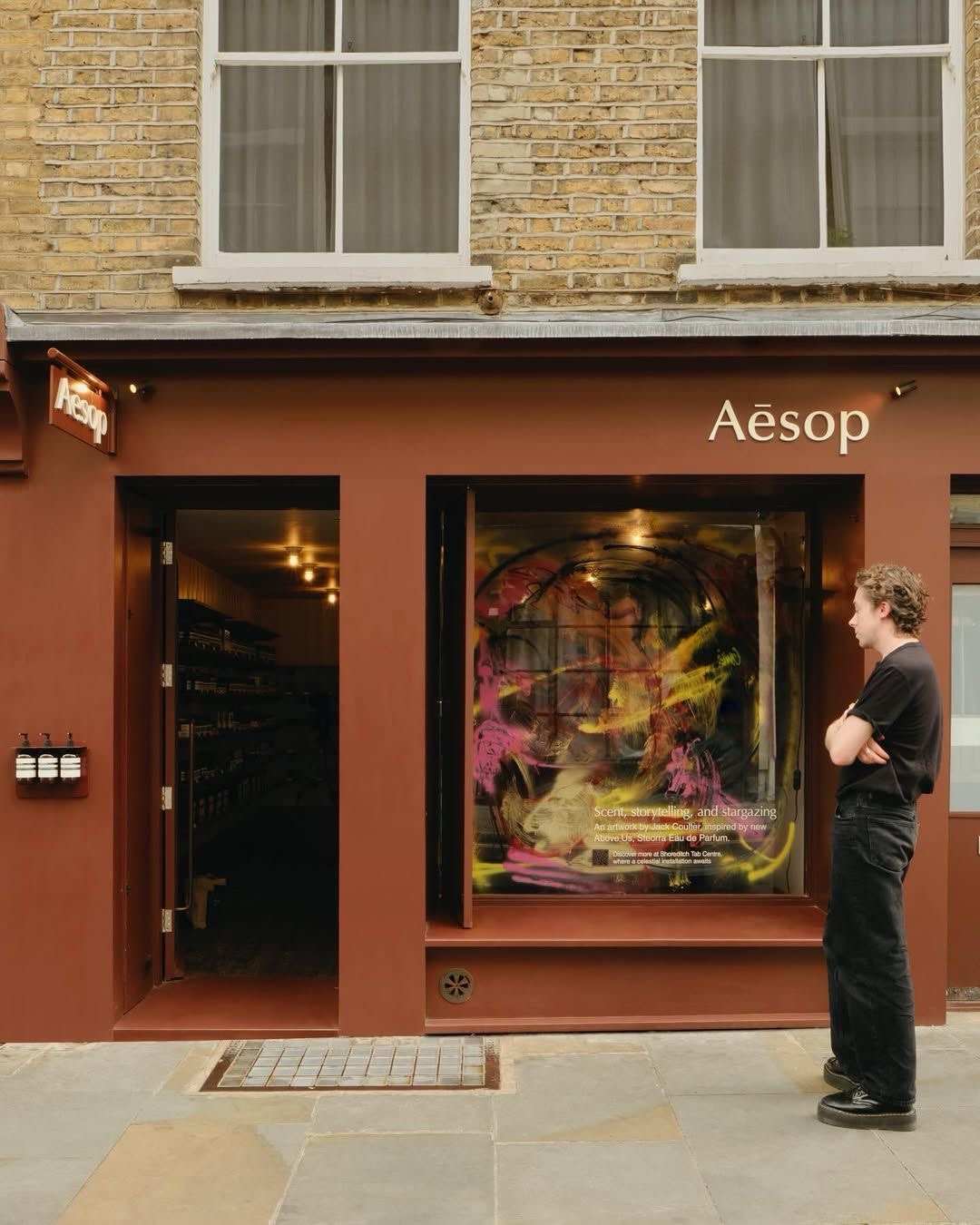 Кирпично-красный фасад Aesop и прохожий у витрины с картиной
