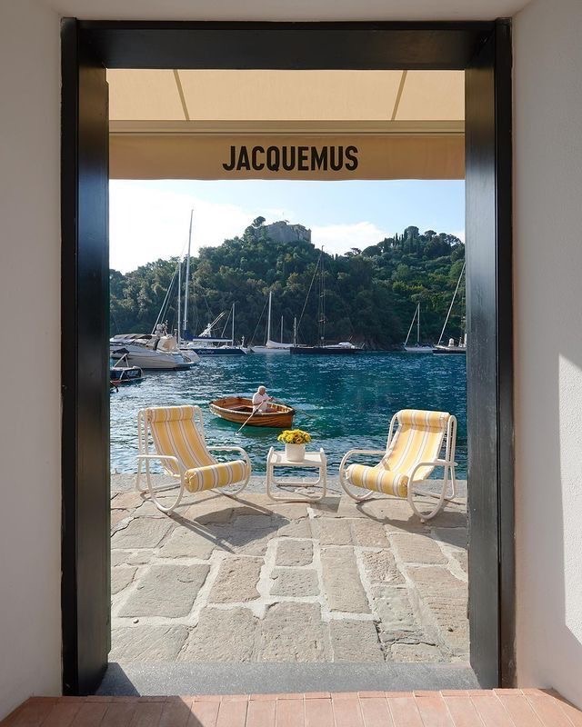 Дверной проём с вывеской Jacquemus, за ним набережная, лодка и два полосатых кресла